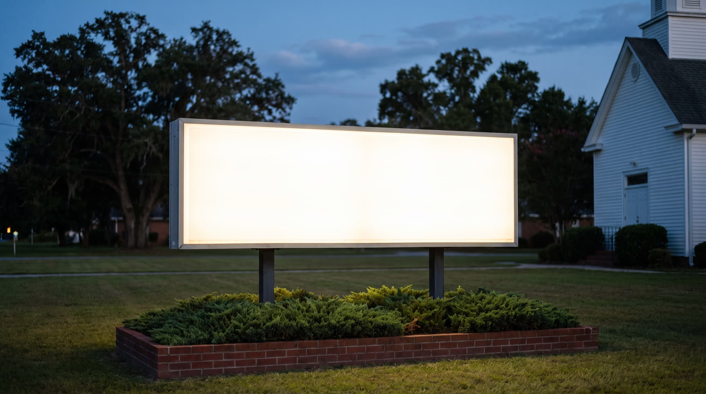 A lit roadside sign on a church lawn at dusk, a white clapboard church and oak trees behind it
