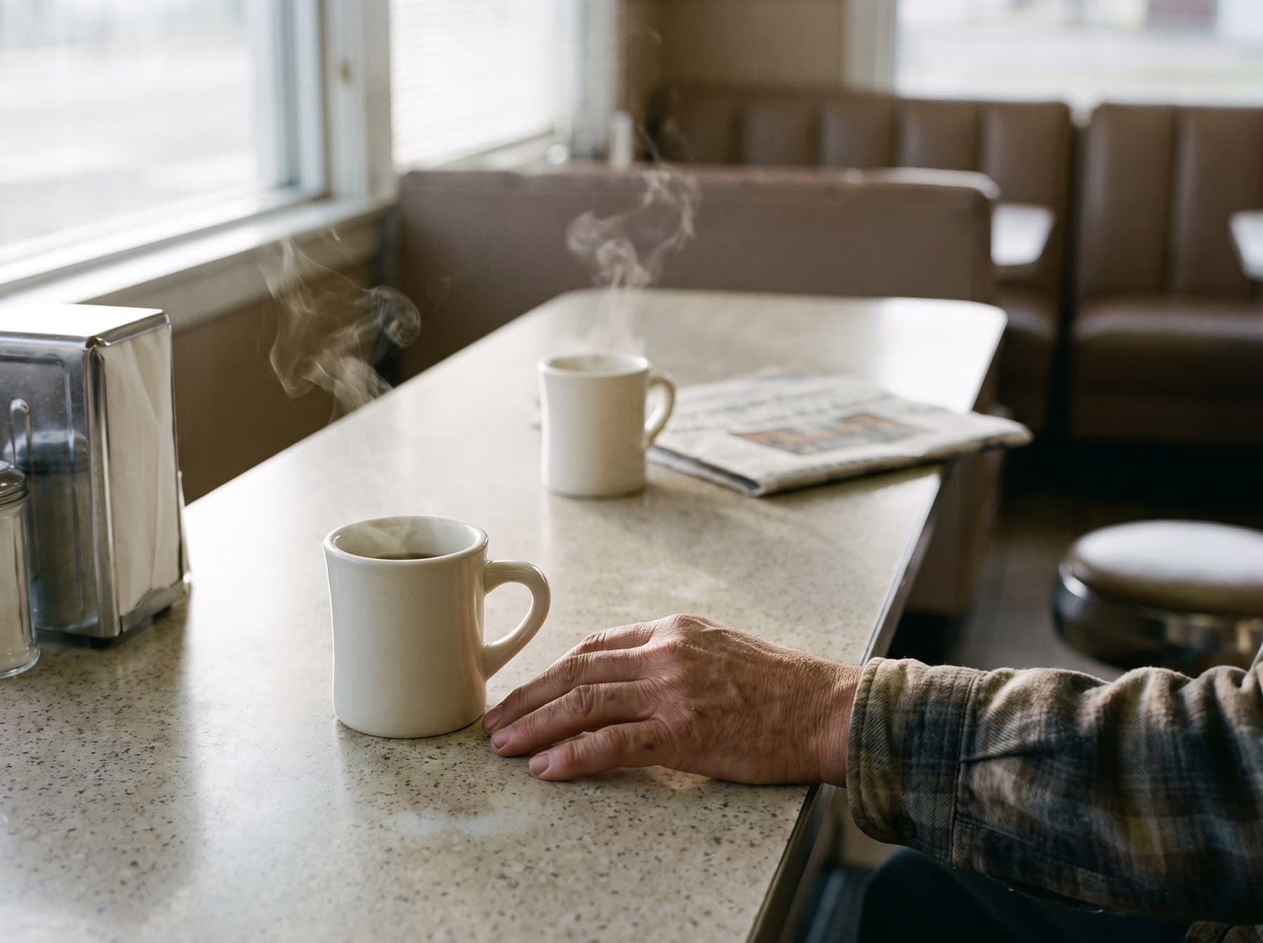 Two white diner mugs of coffee on a worn counter at dawn, a man's hand beside one, the other cup waiting